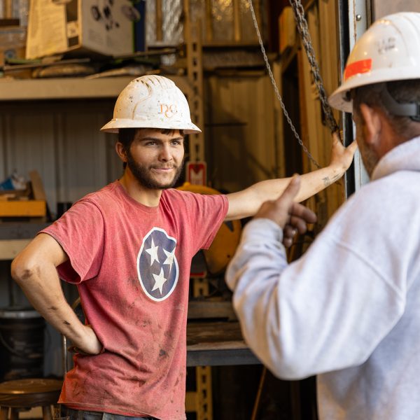 Two men in a warehouse wearing protective helmets talking to one another.